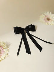 Black satin silk ribbon hair bow with double bow and long tail, styled on a beige background with flowers. Elegant hair accessory for girls and teens.