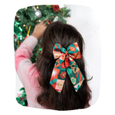 Girl wearing vibrant Christmas hair bow with festive patterns while decorating a Christmas tree. Perfect holiday accessory for girls aged 3 to 12.