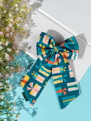 Christmas Gift Hair Bow in green with multicolor gift box prints, displayed on a white textured surface with floral decor.