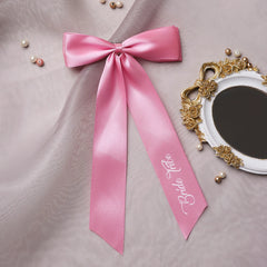 Pink satin Bride Tribe hair bow displayed on soft fabric with pearls, perfect for wedding decor or bridesmaid gifts.