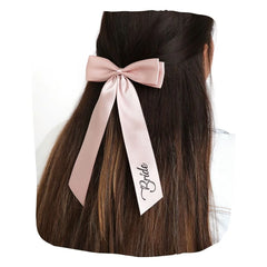 Blush satin hair bow with 'Bride' script, styled on long brunette hair. Ideal bridal accessory for weddings and special occasions.
