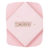 SORRY Clip - Gold Glitter Hairpin for Kids displayed on a pink geometric background. Sparkling rhinestones form the word 'SORRY' in bold letters.