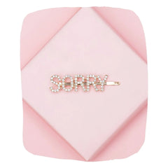 SORRY Clip - Gold Glitter Hairpin for Kids displayed on a pink geometric background. Sparkling rhinestones form the word 'SORRY' in bold letters.
