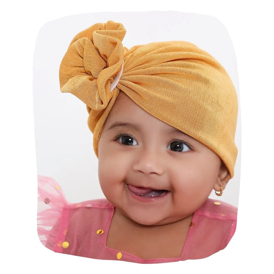 Shiny mustard baby turban cap made of soft cotton, worn by a smiling baby girl. Ideal for newborns and toddlers, perfect for everyday wear.