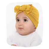 Cotton baby turban in mustard yellow with bow knot, worn by a baby girl. Soft, breathable fabric ideal for newborns and toddlers up to 2 years.
