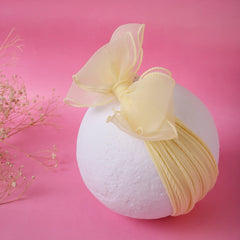 Soft and stretchable yellow baby cotton headband with double layered bow, displayed on a white sphere against a pink background.