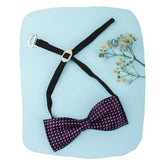 Boys purple satin bow tie with printed dots, adjustable strap and clip-on design, laid flat on blue background with floral accent.