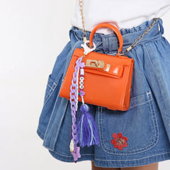 Close-up of orange handbag with beaded bag charm for kids, showing purple chain, tassel, and personalized beads. Ideal for girls' backpacks or purses.