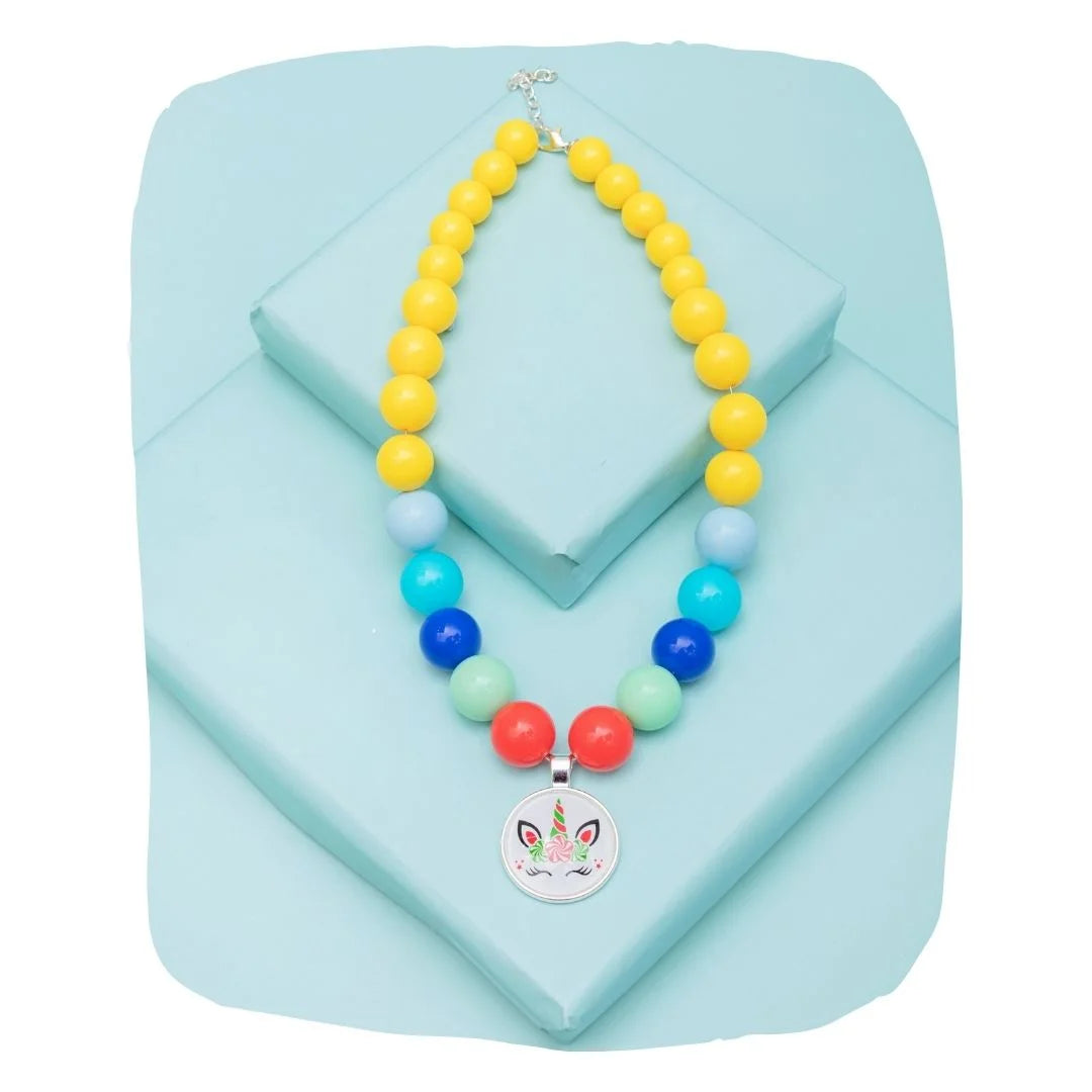 Unicorn necklace for kids with pastel beads in yellow, blue, and red, displayed on a light blue platform. Ideal magical jewelry gift.