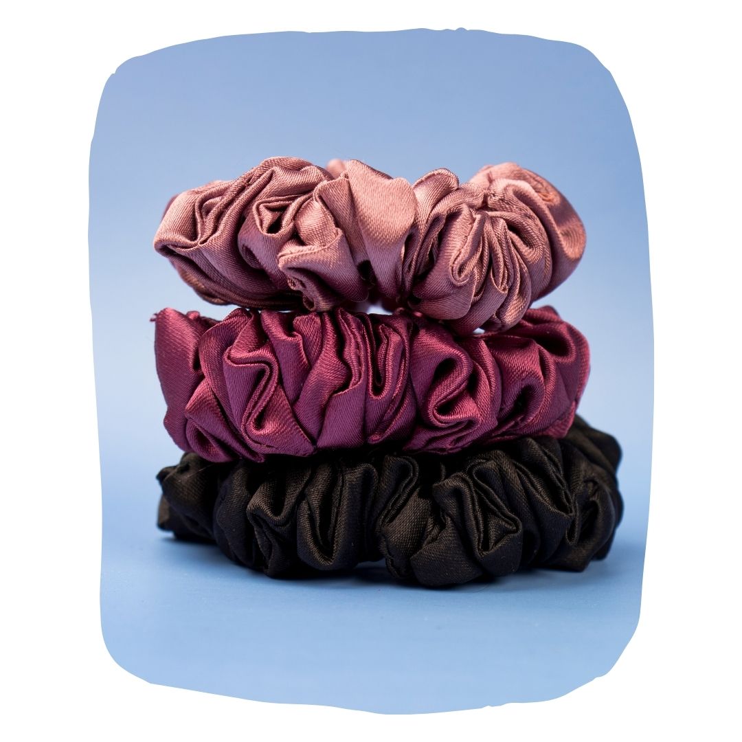 Set of three silk satin scrunchies in pink, burgundy, and black on blue background. Damage-free hold for all hair types, ideal for daily styling.