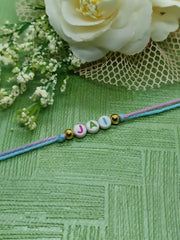 Personalized name Rakhi with 'JAI' beads on pastel thread, styled with white flowers and green textured background for Raksha Bandhan 2025.