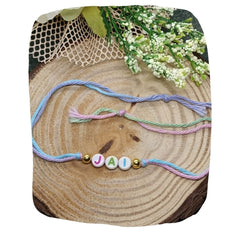 Name Rakhi gift for brother featuring multicolor thread with 'JAI' letter beads and gold accents, displayed on rustic wood with floral decor.