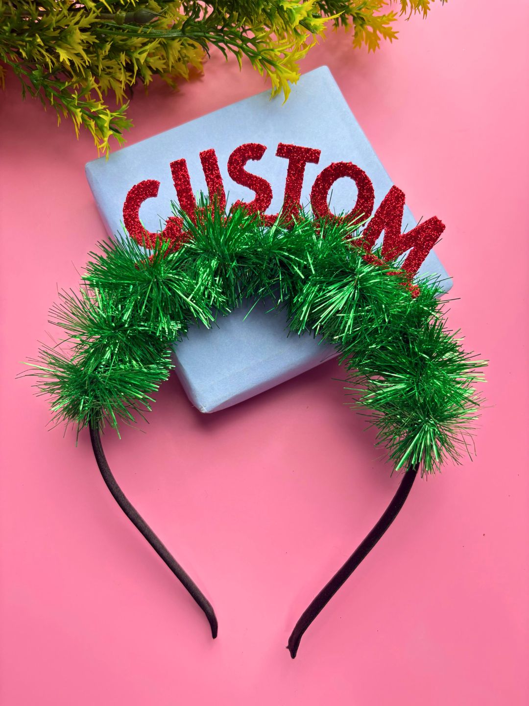 Secure and festive Christmas Glitter Hairband for Girls with green tinsel and red CUSTOM text, styled on a pink background with gift box.
