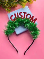 Secure and festive Christmas Glitter Hairband for Girls with green tinsel and red CUSTOM text, styled on a pink background with gift box.