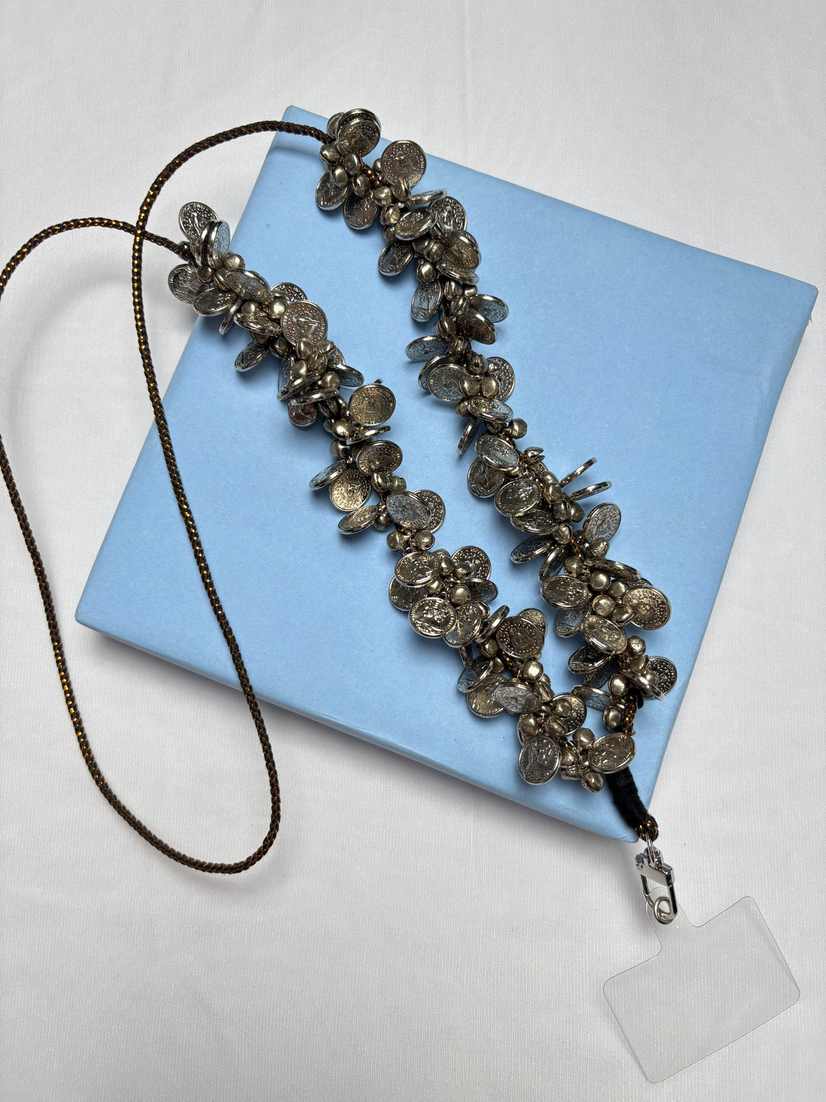 Arendelle Coins Sling - Navratri Style displayed on a blue box, featuring intricate silver coin detailing for festive phone carrying.